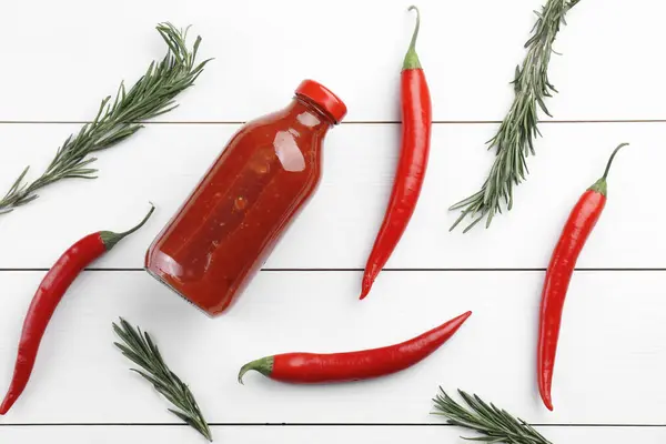 Spicy chili sauce in bottle, peppers and rosemary on white wooden table, flat lay