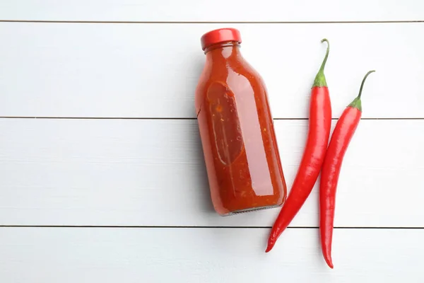 Spicy chili sauce in bottle and peppers on white wooden table, flat lay. Space for text