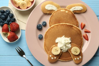 Çocuklar için yaratıcı hizmet. Kreplerden, böğürtlenlerden, kremadan ve muzdan yapılmış, açık mavi ahşap masadaki şirin tavşancıklı tabak.