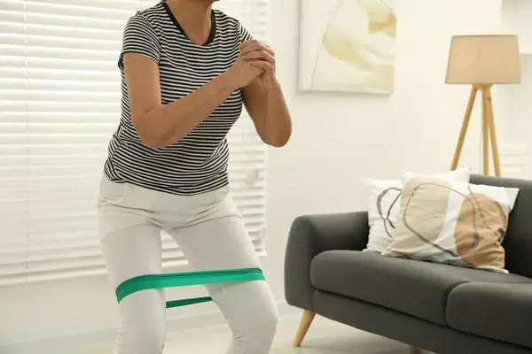 Senior woman doing squats with fitness elastic band at home, closeup. Space for text