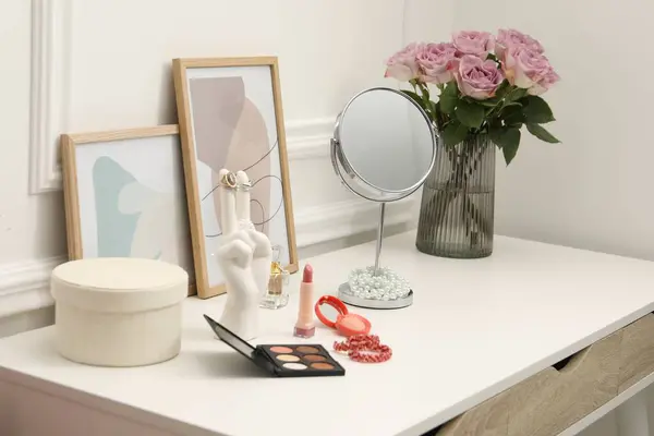 Mirror, cosmetic products, perfume and vase with pink roses on white dressing table in makeup room