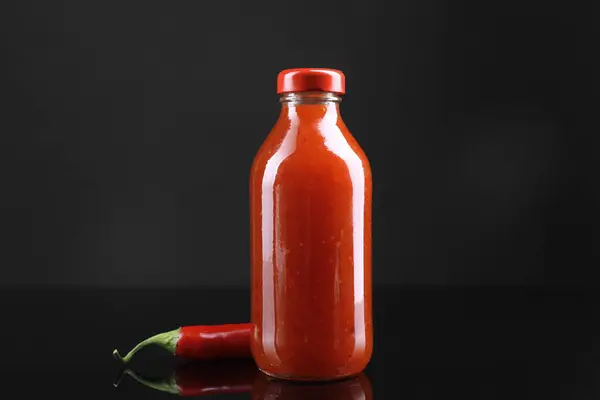 Spicy chili sauce in bottle and pepper against dark background