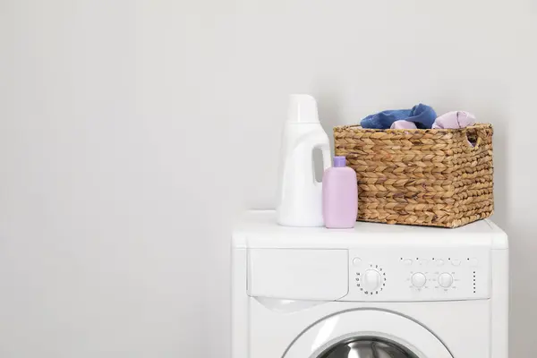 Baby clothes in wicker basket and laundry detergents on washing machine near light wall, space for text