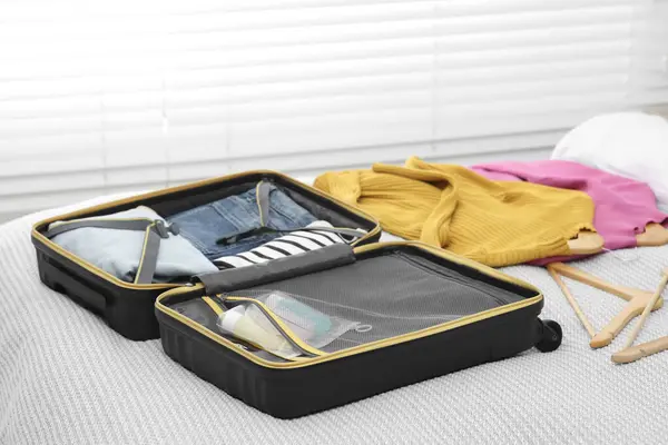 Plastic bag of cosmetic travel kit in suitcase and clothes on bed indoors. Bath accessories