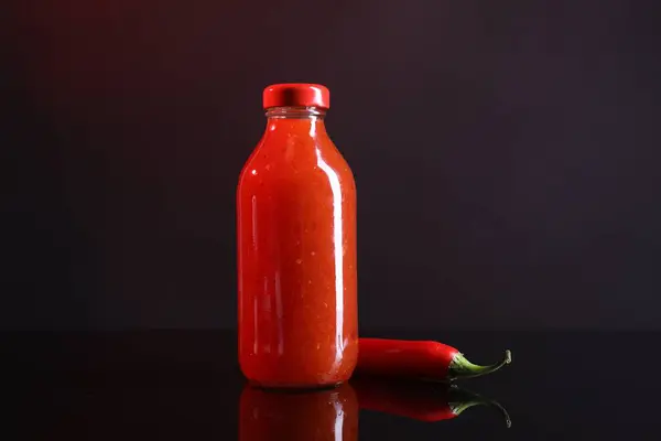 Spicy chili sauce in bottle and pepper against dark background