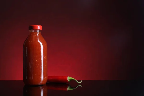 Spicy chili sauce in bottle and pepper against dark background, space for text