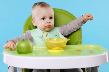 Küçük şirin bebek, açık mavi arka planda, yüksek sandalyede sağlıklı yiyecekler yiyor.