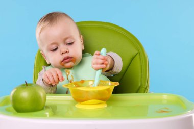 Küçük şirin bebek, açık mavi arka planda, yüksek sandalyede sağlıklı yiyecekler yiyor.