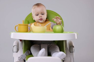 Gri arka planda, yüksek sandalyede sağlıklı yiyecekler yiyen şirin bir bebek.