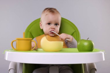 Gri arka planda, yüksek sandalyede sağlıklı yiyecekler yiyen şirin bir bebek.