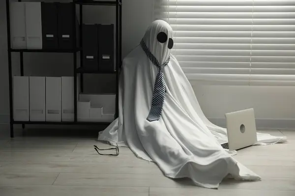 Overworked ghost. Man in white sheet with laptop on floor in office