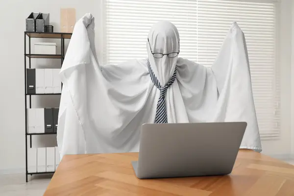 Overworked ghost. Man covered with white sheet using laptop at wooden table in office