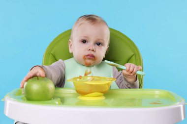 Küçük şirin bebek, açık mavi arka planda, yüksek sandalyede sağlıklı yiyecekler yiyor.