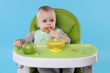 Küçük şirin bebek, açık mavi arka planda, yüksek sandalyede sağlıklı yiyecekler yiyor.