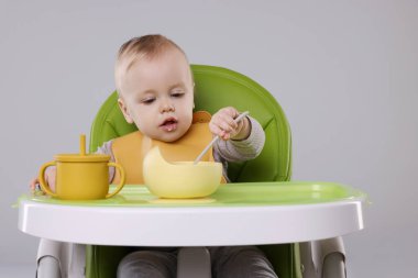 Gri arka planda, yüksek sandalyede sağlıklı yiyecekler yiyen şirin bir bebek.