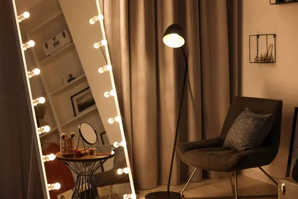 Makeup room. Stylish mirror with light bulbs, beauty products on table and armchair indoors
