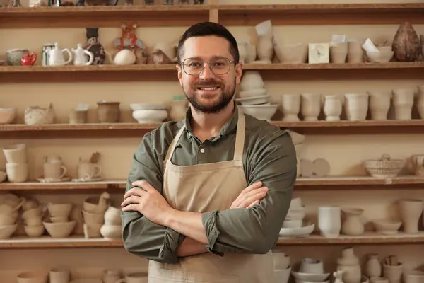 Portrait of smiling craftsman near shelf with different pottery in workshop