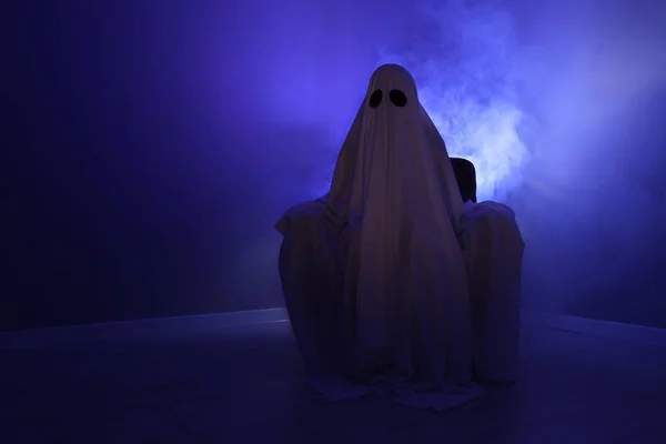 Creepy ghost. Woman covered with sheet sitting in armchair in blue light, space for text