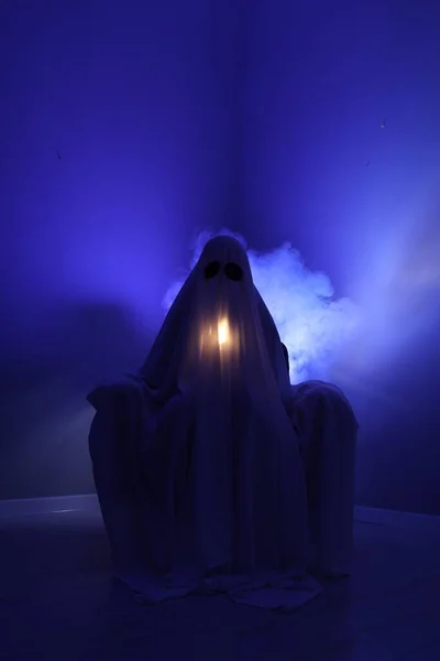 Creepy ghost. Woman covered with sheet in armchair in blue light