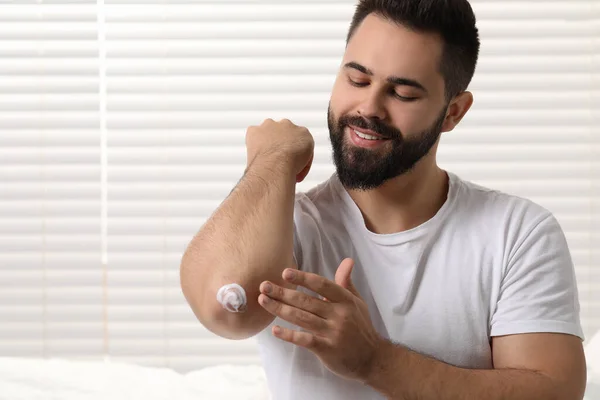 Man with dry skin applying cream onto his elbow indoors, space for text