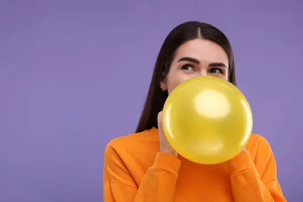 Woman inflating yellow balloon on purple background, space for text