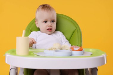 Turuncu arka planda, yüksek sandalyede sağlıklı yiyecekleri olan şirin bir bebek.