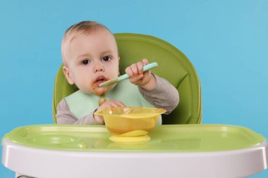 Küçük şirin bebek, açık mavi arka planda, yüksek sandalyede sağlıklı yiyecekler yiyor.