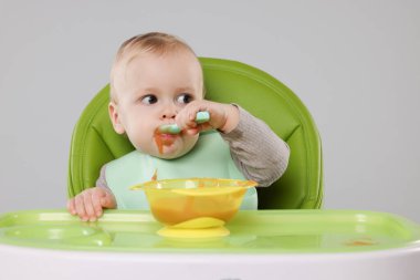 Gri arka planda, yüksek sandalyede sağlıklı yiyecekler yiyen şirin bir bebek.