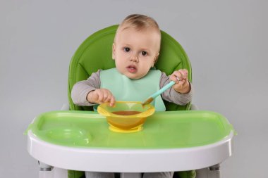 Gri arka planda, yüksek sandalyede sağlıklı yiyecekler yiyen şirin bir bebek.