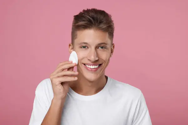 Handsome man cleaning face with cotton pad on pink background