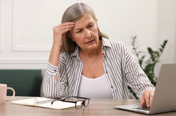 Menopause. Woman suffering from headache while using laptop at wooden table indoors