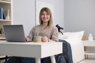 Tekerlekli sandalyedeki kadın bir bardak içkiyle ofisteki masada dizüstü bilgisayar kullanıyor.