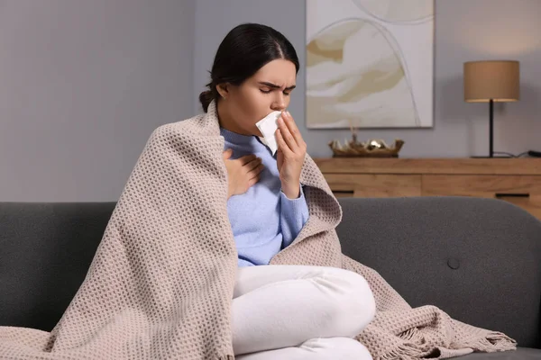 Woman coughing with tissue on sofa at home. Cold symptoms