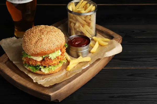 Delicious burger with crispy chicken patty, french fries, sauce and beer on black wooden table