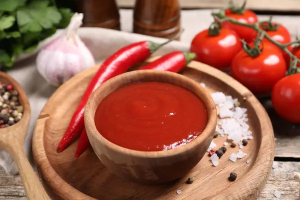 Delicious ketchup in bowl, chili pepper and spices on wooden table, closeup. Tomato sauce