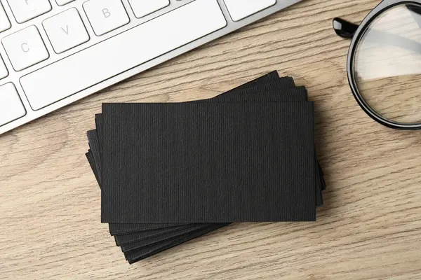 Blank black business cards, computer keyboard and glasses on wooden table, flat lay. Mockup for design