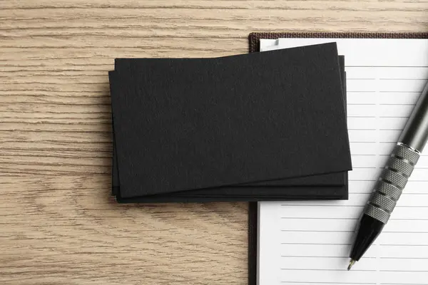 Blank black business cards, notebook and pen on wooden table, top view. Mockup for design