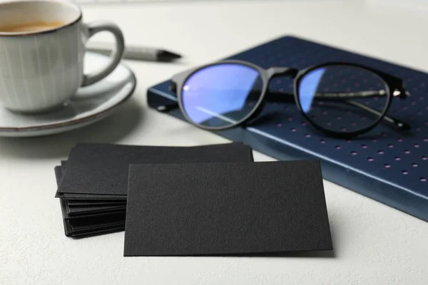 Blank black business cards, notebook, glasses and cup of coffee on white table, closeup. Mockup for design