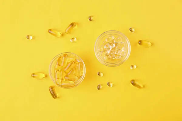 Different vitamin capsules in glass bowls on yellow background, flat lay