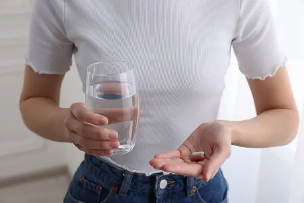 Woman with vitamin pills and glass of water indoors, closeup