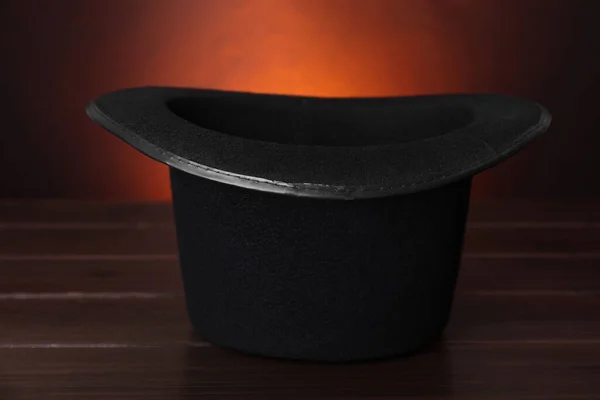 Black magician top hat on wooden table against color background, closeup