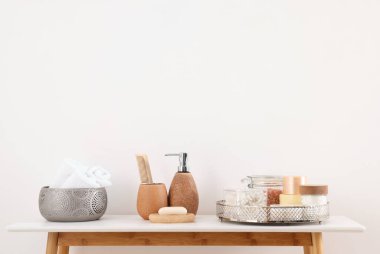 Beyaz duvarın yanındaki masada farklı banyo aksesuarları ve kişisel bakım ürünleri var.