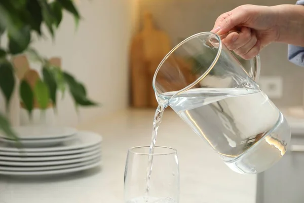 Woman pouring water from jug into glass at white table in kitchen, closeup