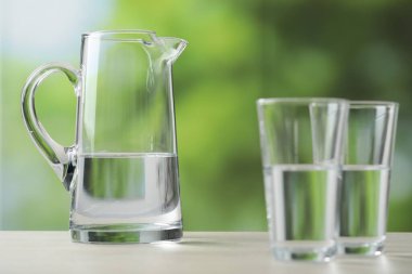 Bulanık yeşil arka planda, temiz su bulunan sürahi ve bardaklar, yakın plan.