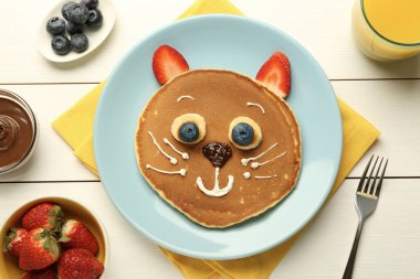 Çocuklar için yaratıcı hizmet. Beyaz ahşap masada krep, böğürtlen, krema, muz ve çikolata ezmesinden yapılmış şirin bir kedi tabağı.