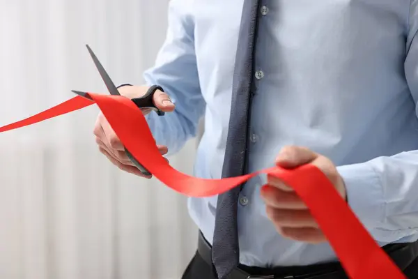 Man cutting red ribbon with scissors indoors, closeup
