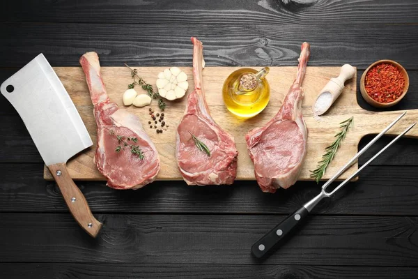 Fresh tomahawk beef cuts, spices and butcher tools on black wooden table, top view