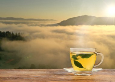 Aromatik yeşil çay cam bardakta, ahşap masada, sisli dağlarda gün doğumunda, metin için yer var.