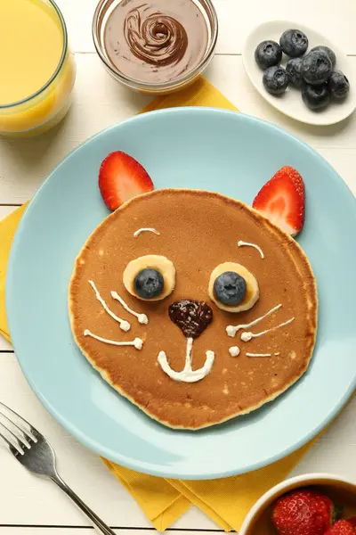 Creative serving for kids. Plate with cute cat made of pancakes, berries, cream, banana and chocolate paste on white wooden table, flat lay