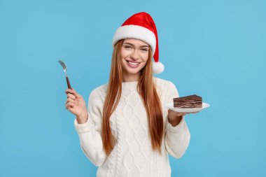 Noel Baba şapkalı genç bir kadın açık mavi arka planda lezzetli bir pastayla